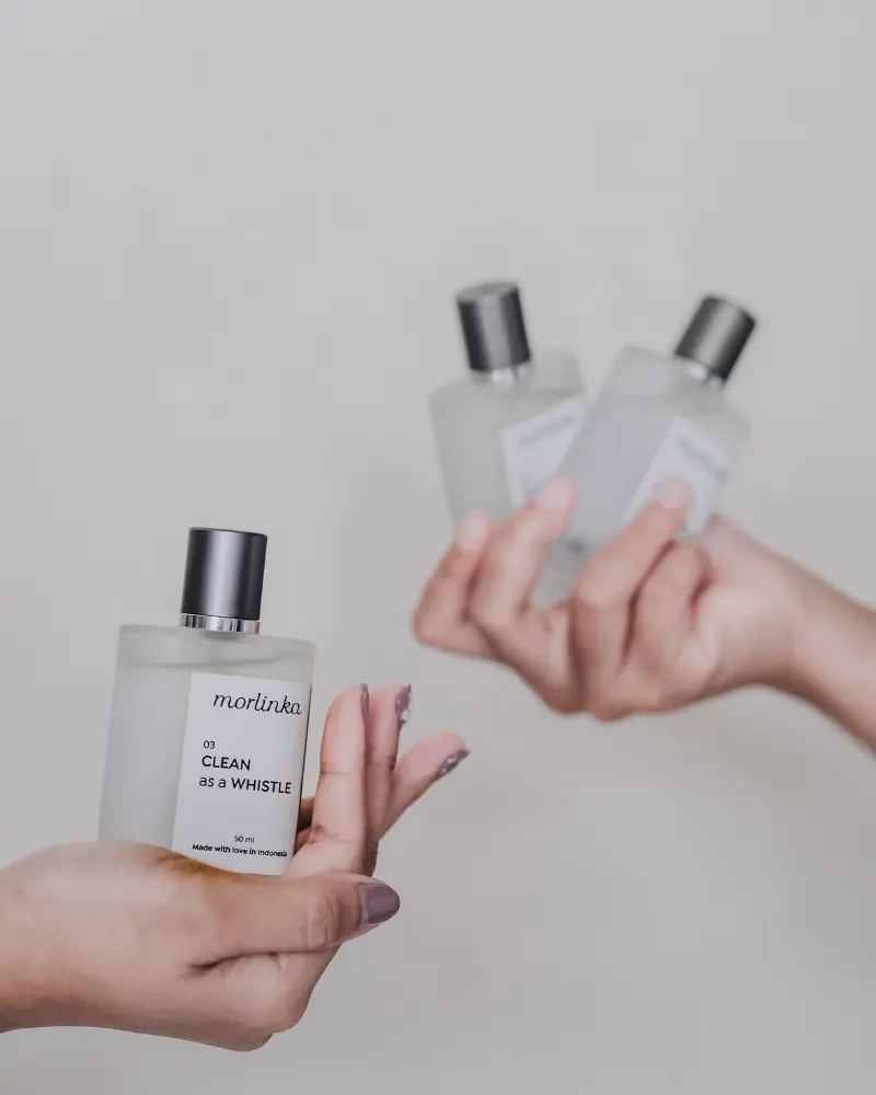 Hands holding Morlinka “Clean as a Whistle” crystal mist bottles, emphasizing modern packaging and a clean lifestyle aesthetic.